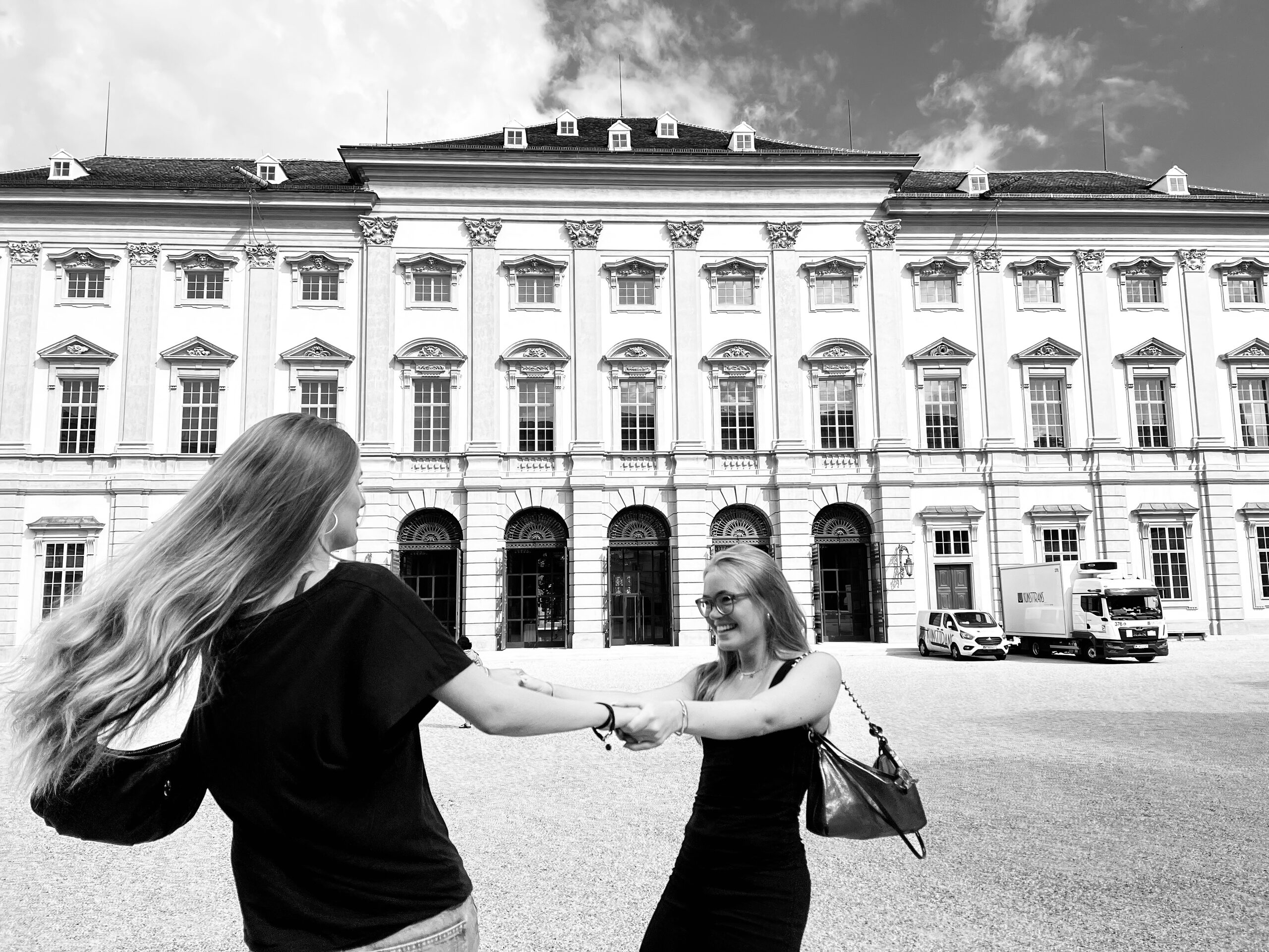 Two women turning hand in hand in front of the long baroque facade of a palace.
